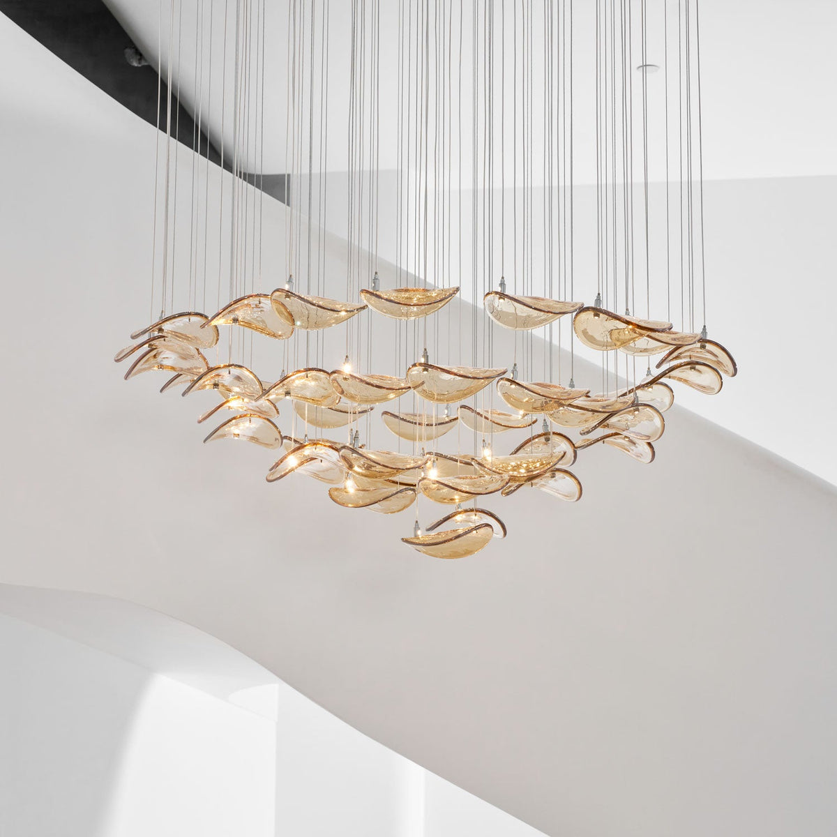 Amber glass leaves in tiered rings, from this Clérachic pendant, hung against the white curve of a staircase.