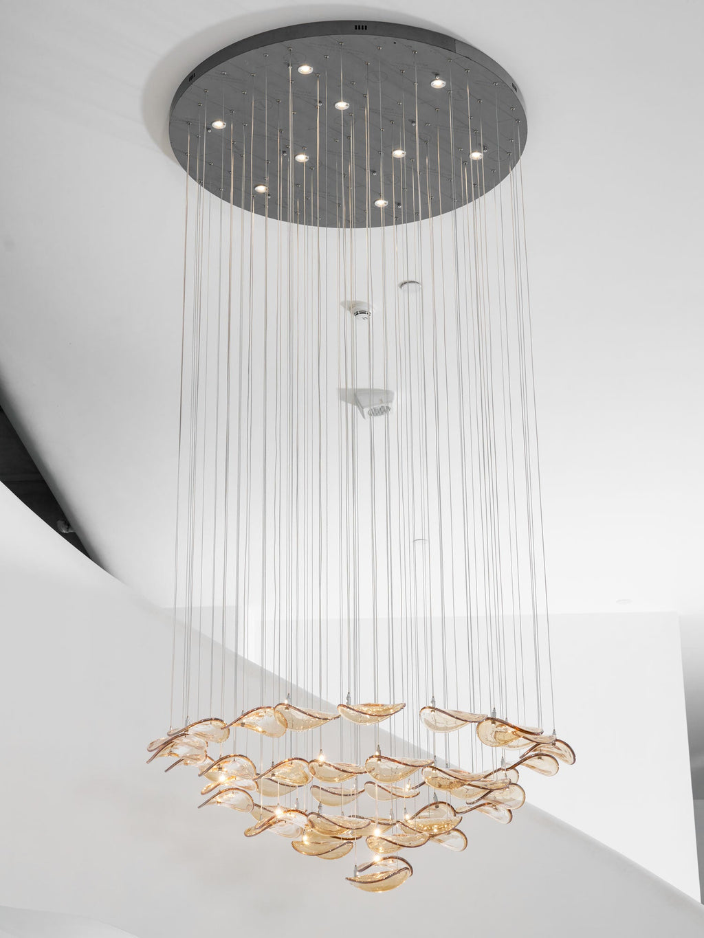 Round canopy with small lights above a long drop of cables, ending in a layered ring of amber glass leaves from Clérachic.