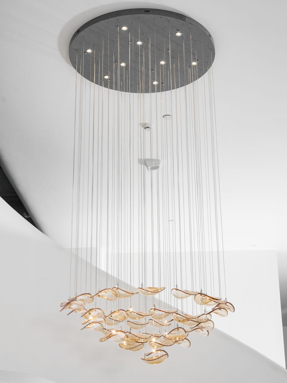 Round canopy with small lights above a long drop of cables, ending in a layered ring of amber glass leaves from Clérachic.