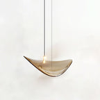 Single amber glass leaf with a small light on its upper face, hung on a dark cable, from this Clérachic pendant, on a white backdrop.