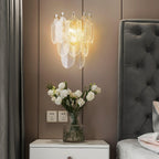 Lit Clérachic glass-leaf wall light above a white bedside table with white roses, against a taupe wall and a grey tufted headboard.