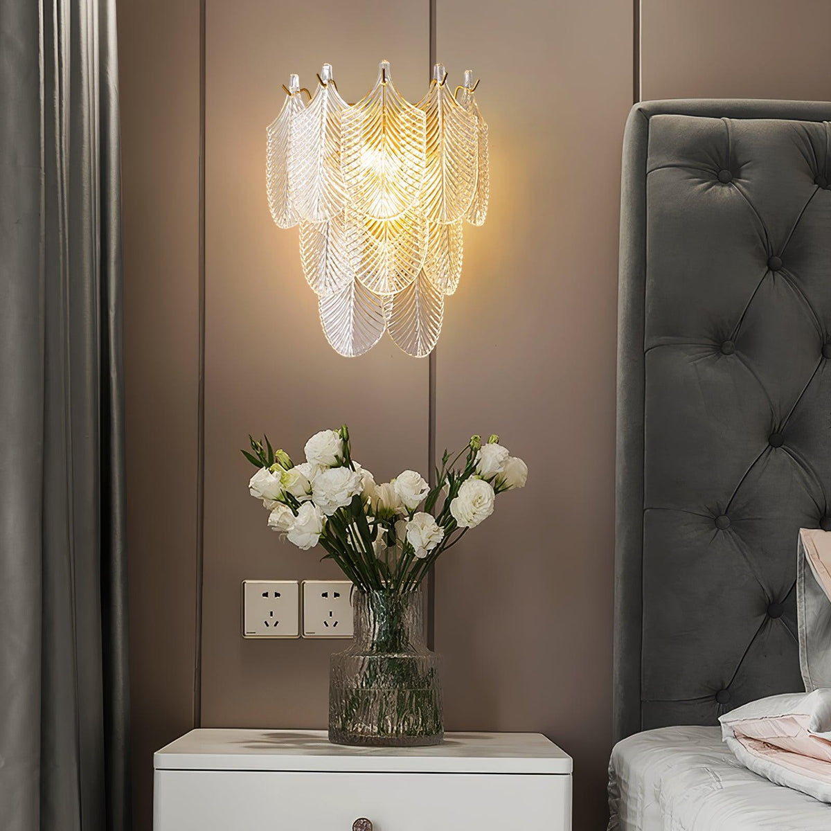 Lit Clérachic glass-leaf wall light above a white bedside table with white roses, against a taupe wall and a grey tufted headboard.