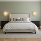 Two Clérachic wall lights either side of an upholstered bed on a sage-green wall, with dark bedside tables and white bedding.