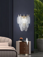 Clérachic glass-leaf wall light lit cool-white on a navy wall, above a bedside chest of drawers with a white figurine.