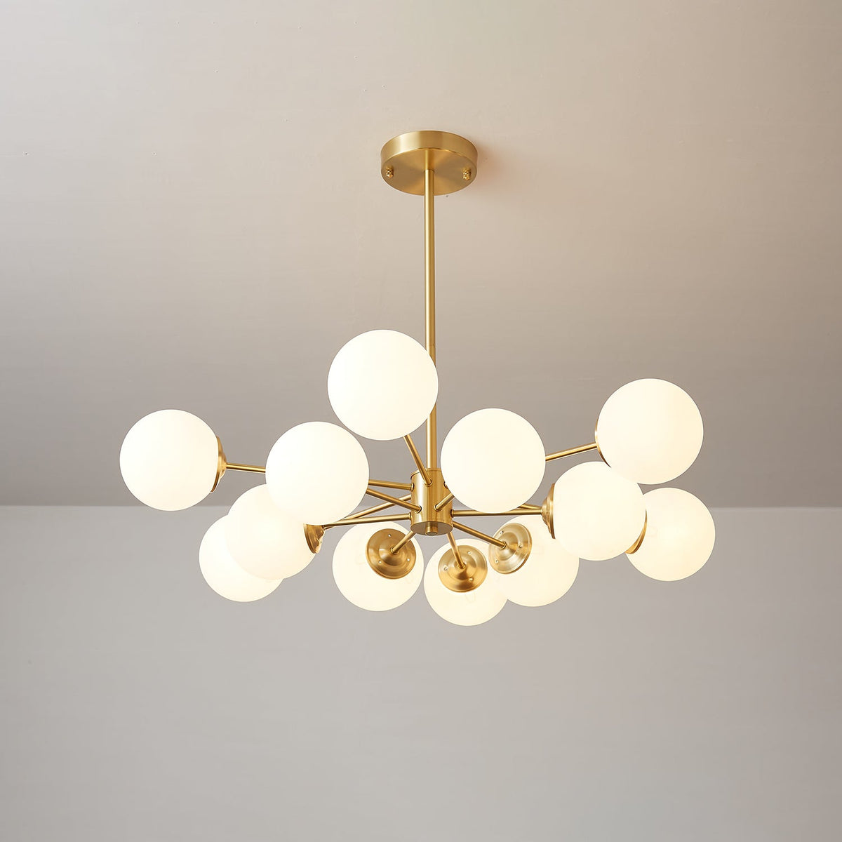 A Clérachic chandelier with lit white glass orbs at varied heights around a central brass stem, on a warm grey backdrop.