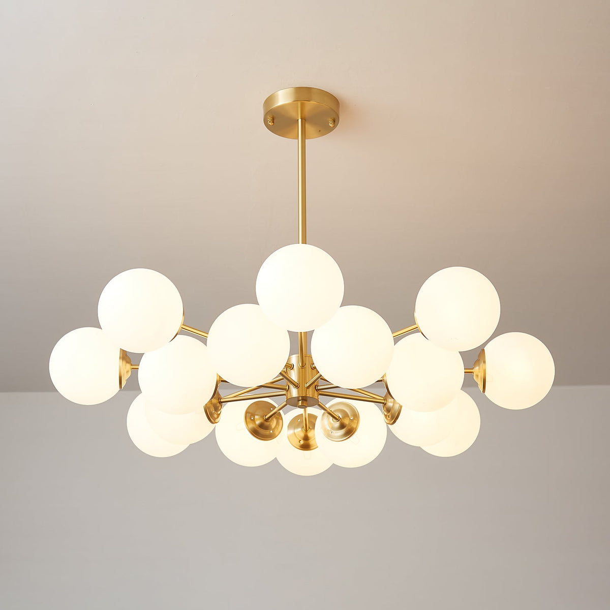 A Clérachic chandelier with a full cluster of lit orbs, the brass drop rod and canopy above, on a warm taupe wall.