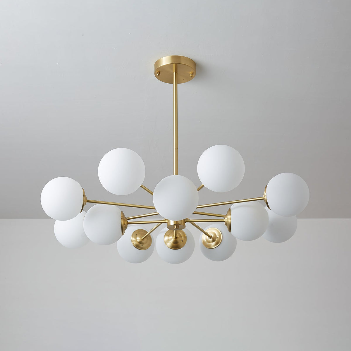 Unlit white orbs on slim brass arms, a Clérachic chandelier seen against a pale grey wall.