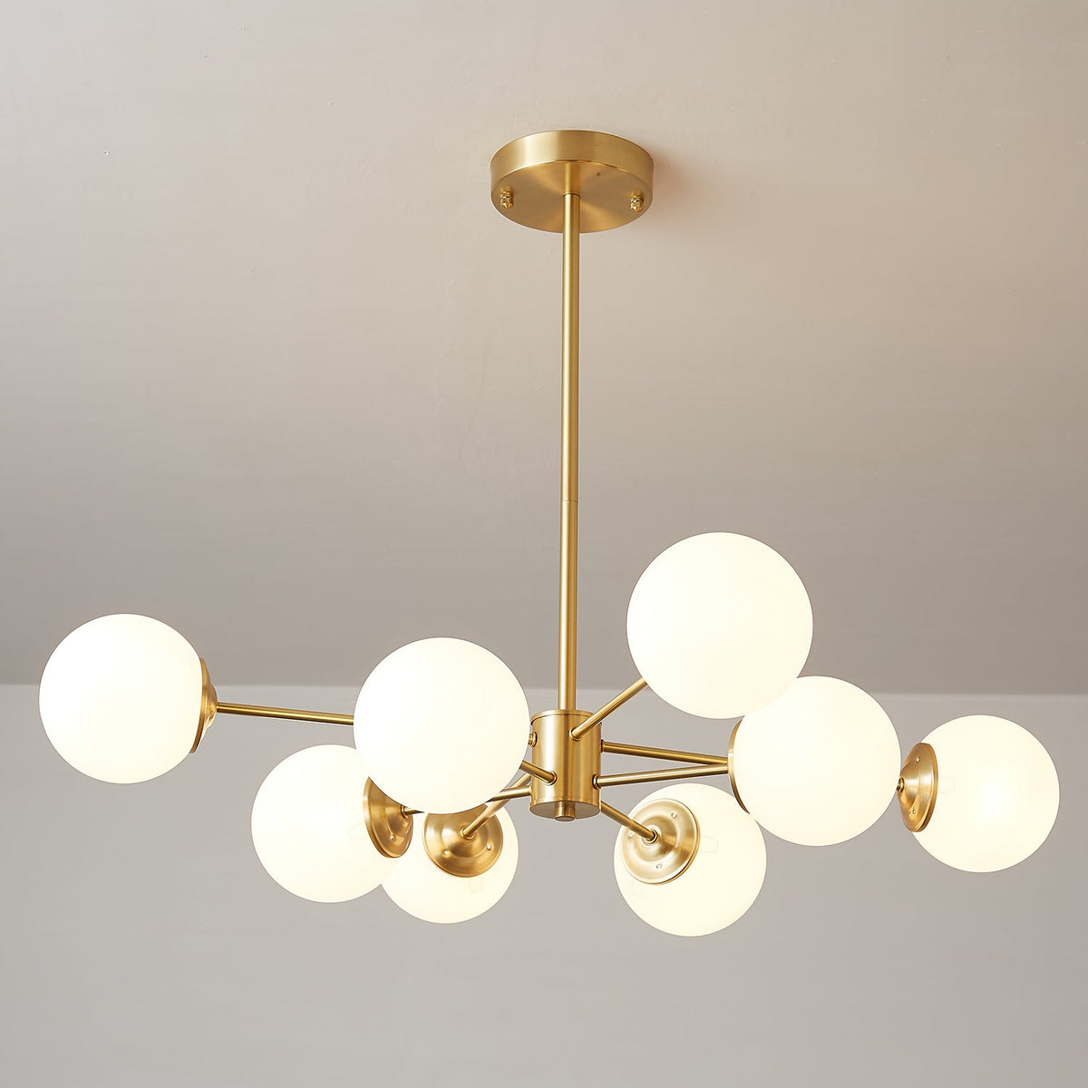 Clérachic chandelier with eight lit orbs spaced wide on long brass arms, on a soft grey backdrop.