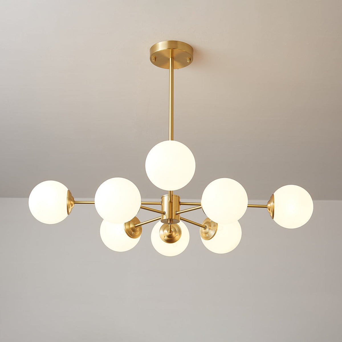 Wide-armed Clérachic chandelier with lit white orbs and a central brass hub, against a warm taupe wall.