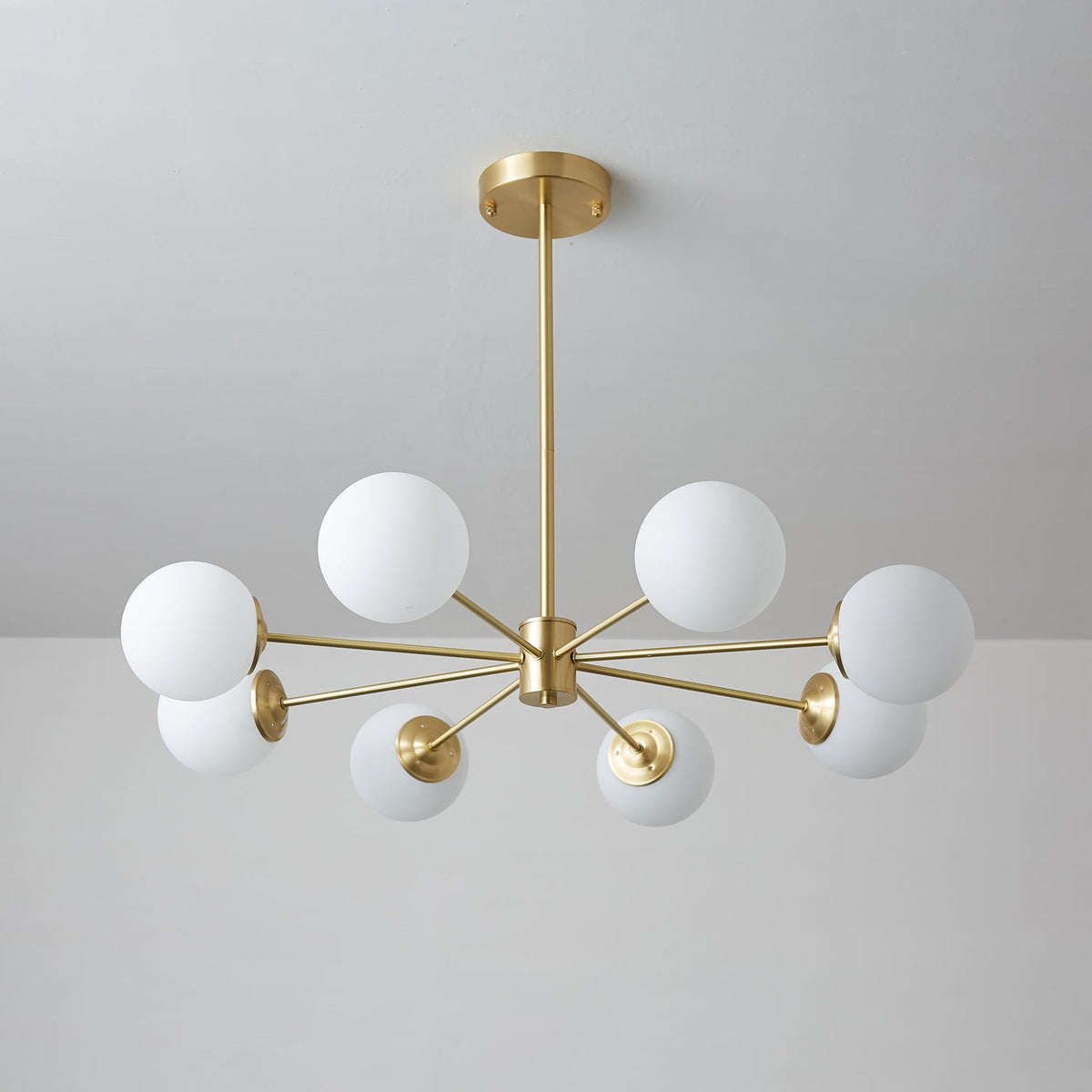 Unlit Clérachic chandelier with matt white orbs on long, slim brass arms, set against a cool grey wall.