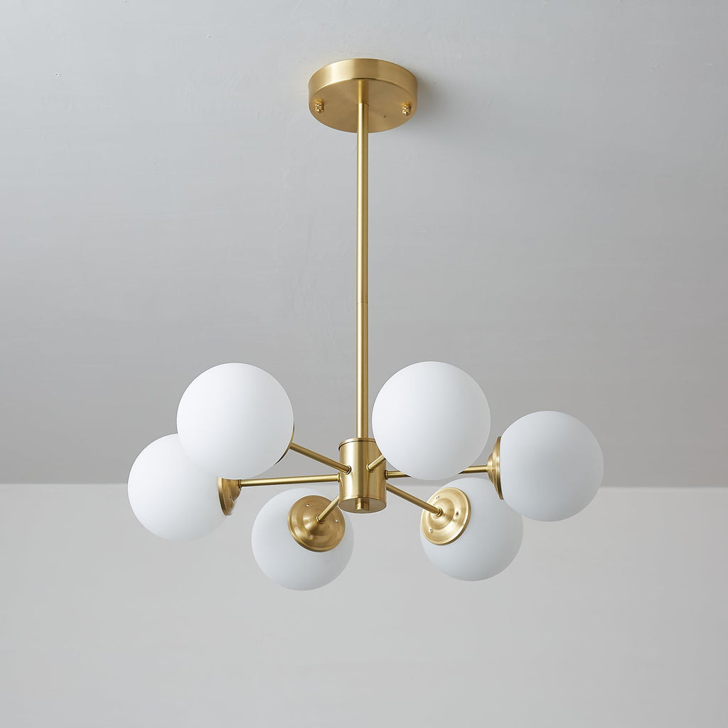 Unlit Clérachic chandelier with larger matt white orbs on short brass arms, against a cool grey wall.