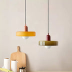 Beside a wooden chopping board on a pale wall, an orange disc pendant with a pink cap and an olive one with a red cap, from Clérachic.