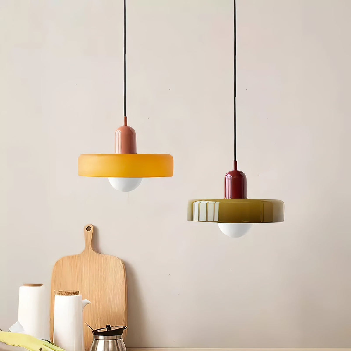 Beside a wooden chopping board on a pale wall, an orange disc pendant with a pink cap and an olive one with a red cap, from Clérachic.