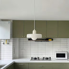 A white disc pendant with a white cap hangs above a kitchen with sage-green cabinets and white tiling, in this Clérachic photograph.
