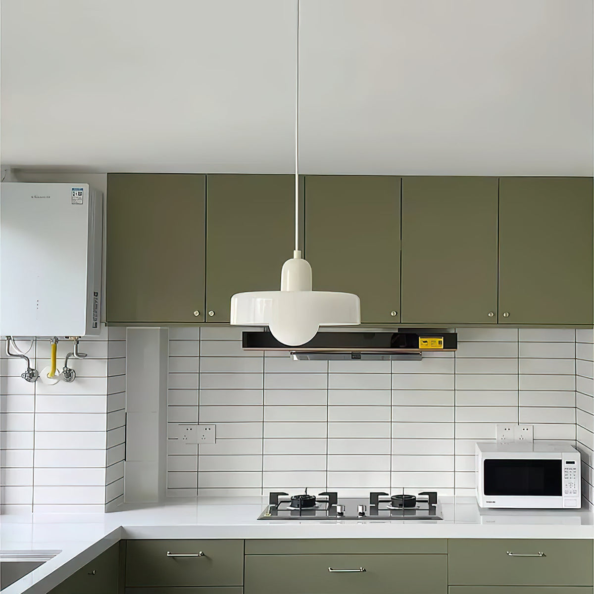 A white disc pendant with a white cap hangs above a kitchen with sage-green cabinets and white tiling, in this Clérachic photograph.