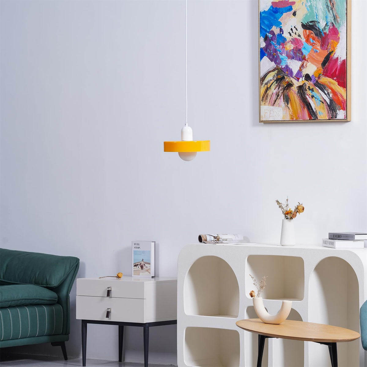 An orange disc pendant with a white cap hangs against a pale wall, below a colourful painting and above a teal sofa, from Clérachic.