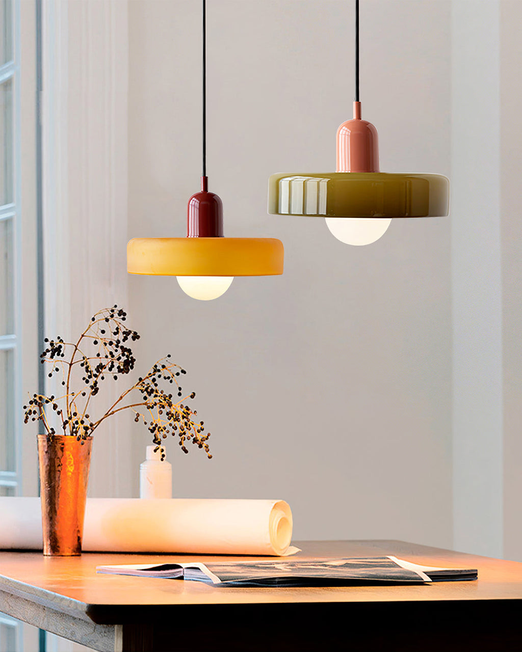 An orange disc with a deep red cap and an olive-green disc with a pink cap hang over a wooden desk by a window, from Clérachic.