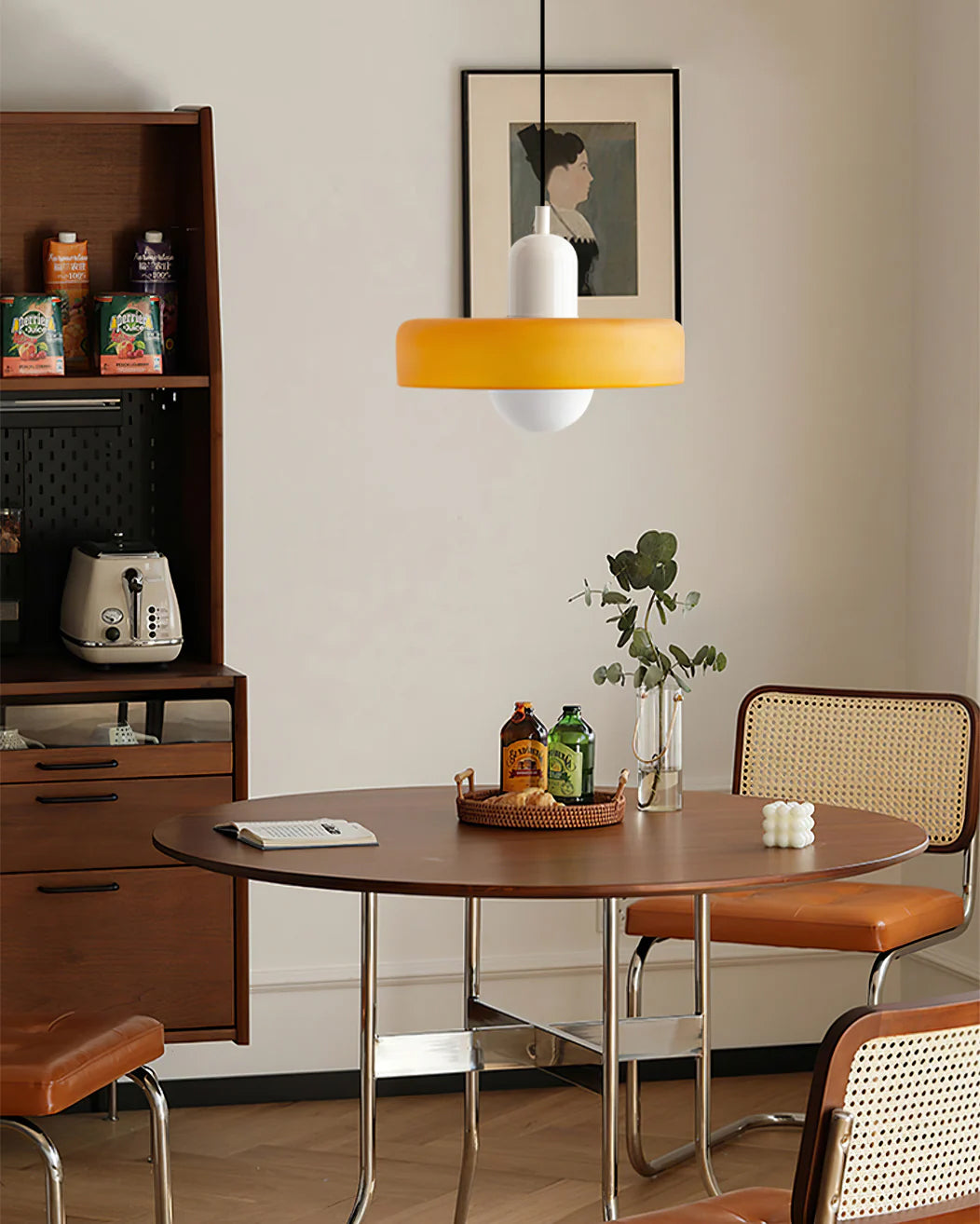 An orange disc pendant with a white cap hangs above a round walnut table with rattan and leather chairs, a Clérachic accent.