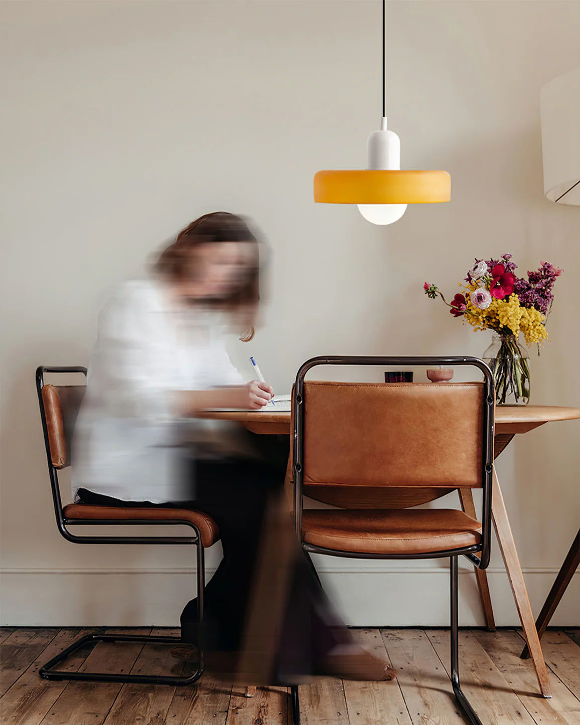 A blurred figure writes at a wooden table under a Clérachic orange disc pendant with a white cap, beside a vase of wildflowers.