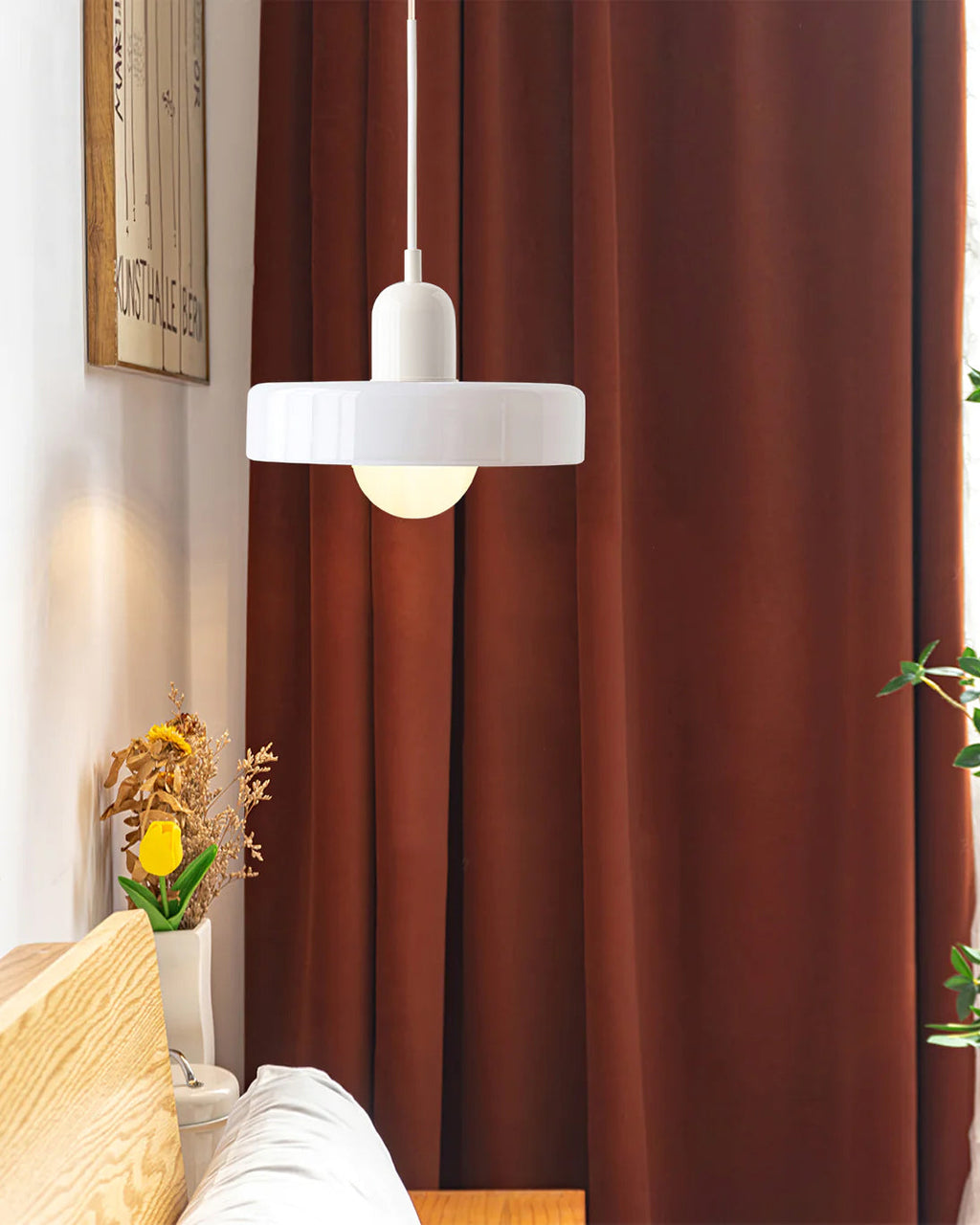 A white disc pendant with a white cap hangs before a rust-red velvet curtain, a Clérachic light in a cosy corner of a room.
