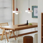 Two white disc pendants with deep red caps hang above a long wooden table with a tea set, with rattan chairs and a blind beyond, from Clérachic.