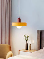 An orange disc pendant with a deep red cap hangs by a bedside with a pink curtain and two small vases, a Clérachic bedroom light.