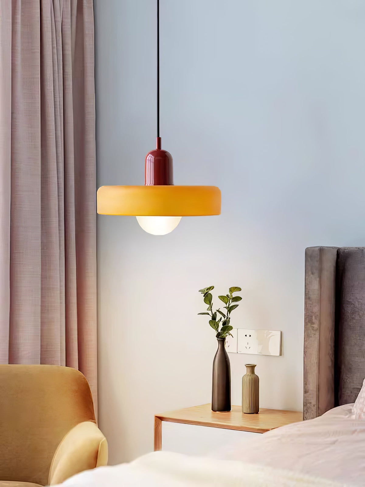 An orange disc pendant with a deep red cap hangs by a bedside with a pink curtain and two small vases, a Clérachic bedroom light.