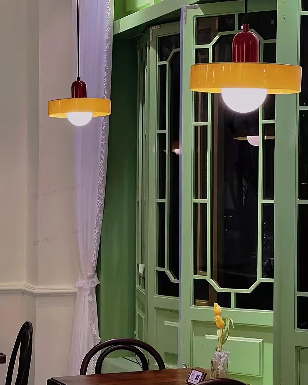 Two orange disc pendants with deep red caps glow in front of a green lattice window, in an evening café scene from Clérachic.