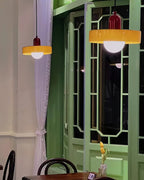 Two orange disc pendants with deep red caps glow in front of a green lattice window, in an evening café scene from Clérachic.