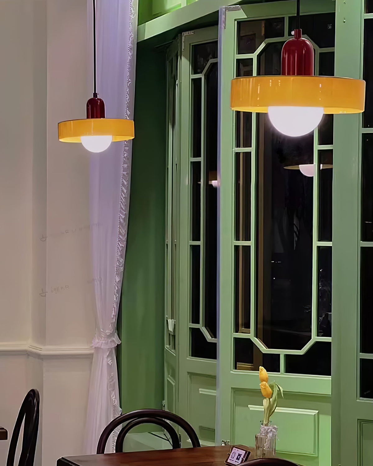 Two orange disc pendants with deep red caps glow in front of a green lattice window, in an evening café scene from Clérachic.