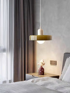 By the bed, a Clérachic olive-green disc pendant with a beige cap glows softly on a white cord, next to a grey curtain and a bedside table.