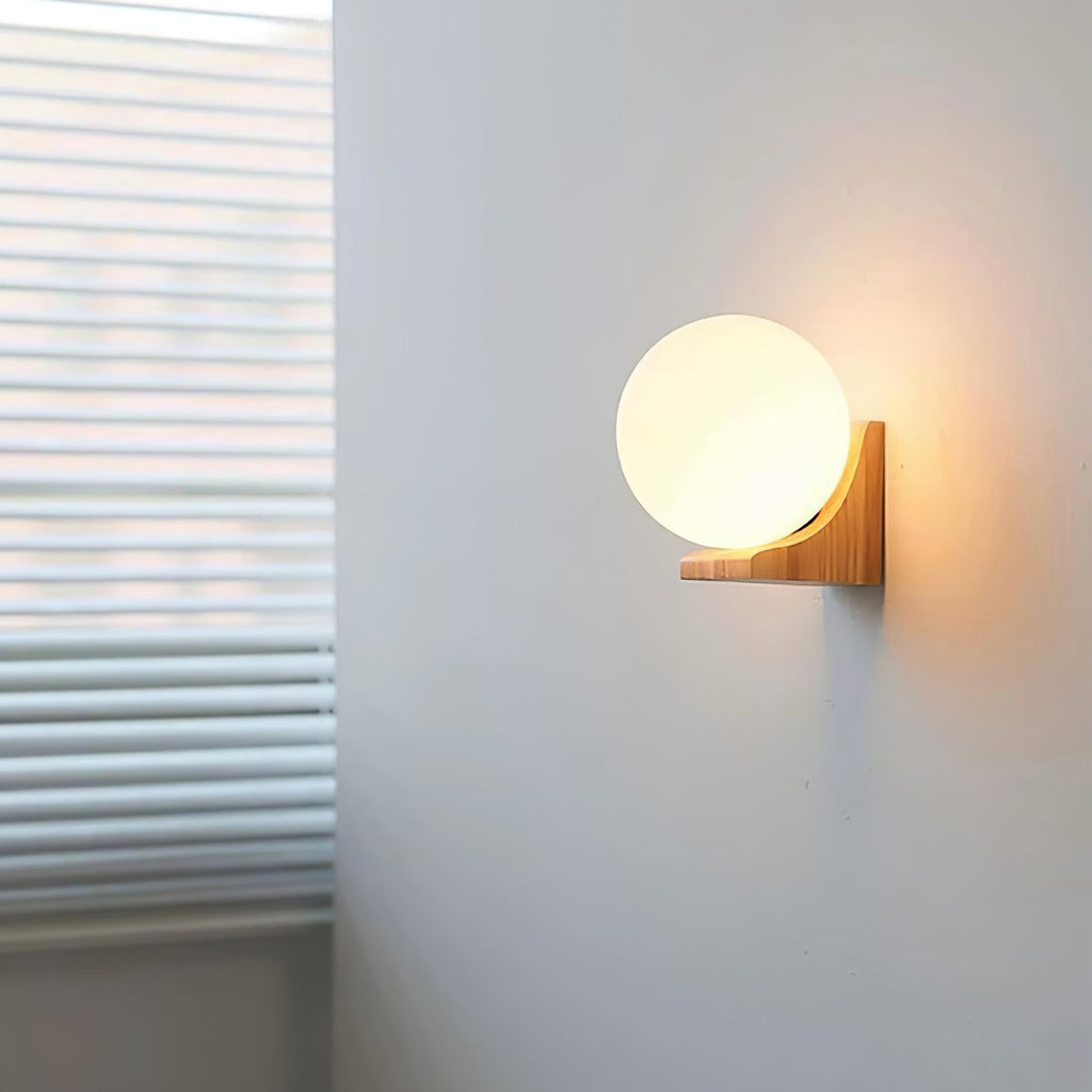 Small lit Clérachic glass orb wall light on a pale grey wall, with window blinds at the left and a warm glow behind the bracket.