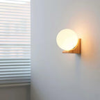 Small lit Clérachic glass orb wall light on a pale grey wall, with window blinds at the left and a warm glow behind the bracket.