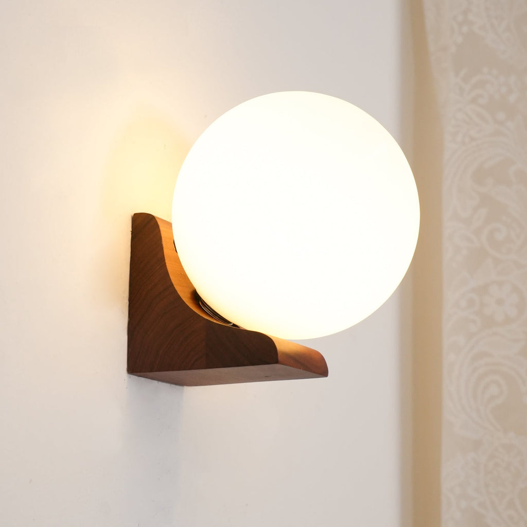 Lit Clérachic orb wall light on a cream wall beside lace curtain, its dark wood bracket supporting the glass from below.