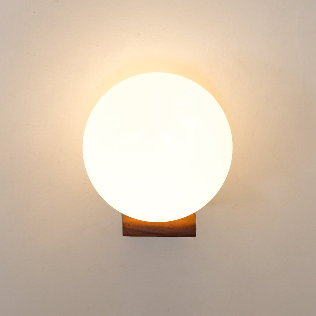 Front view of a warmly lit glass sphere above a short wooden bracket, this Clérachic wall light on a cream wall.