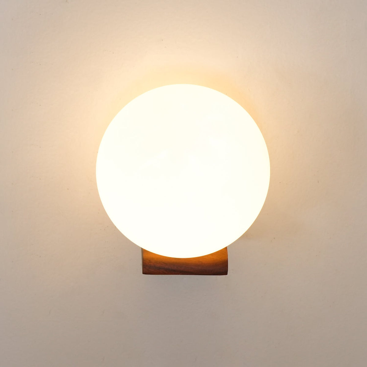 Front view of a warmly lit glass sphere above a short wooden bracket, this Clérachic wall light on a cream wall.