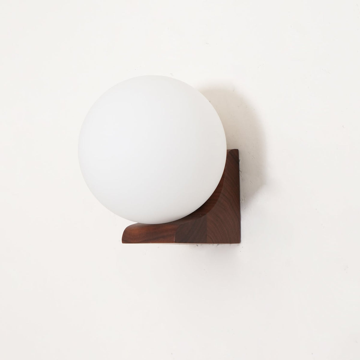Opaque white glass globe resting in a walnut bracket, this Clérachic wall light seen unlit from slightly below.