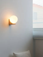 Tall room view of a Clérachic orb wall light glowing beside a window with blinds, with a cushion in the corner below.