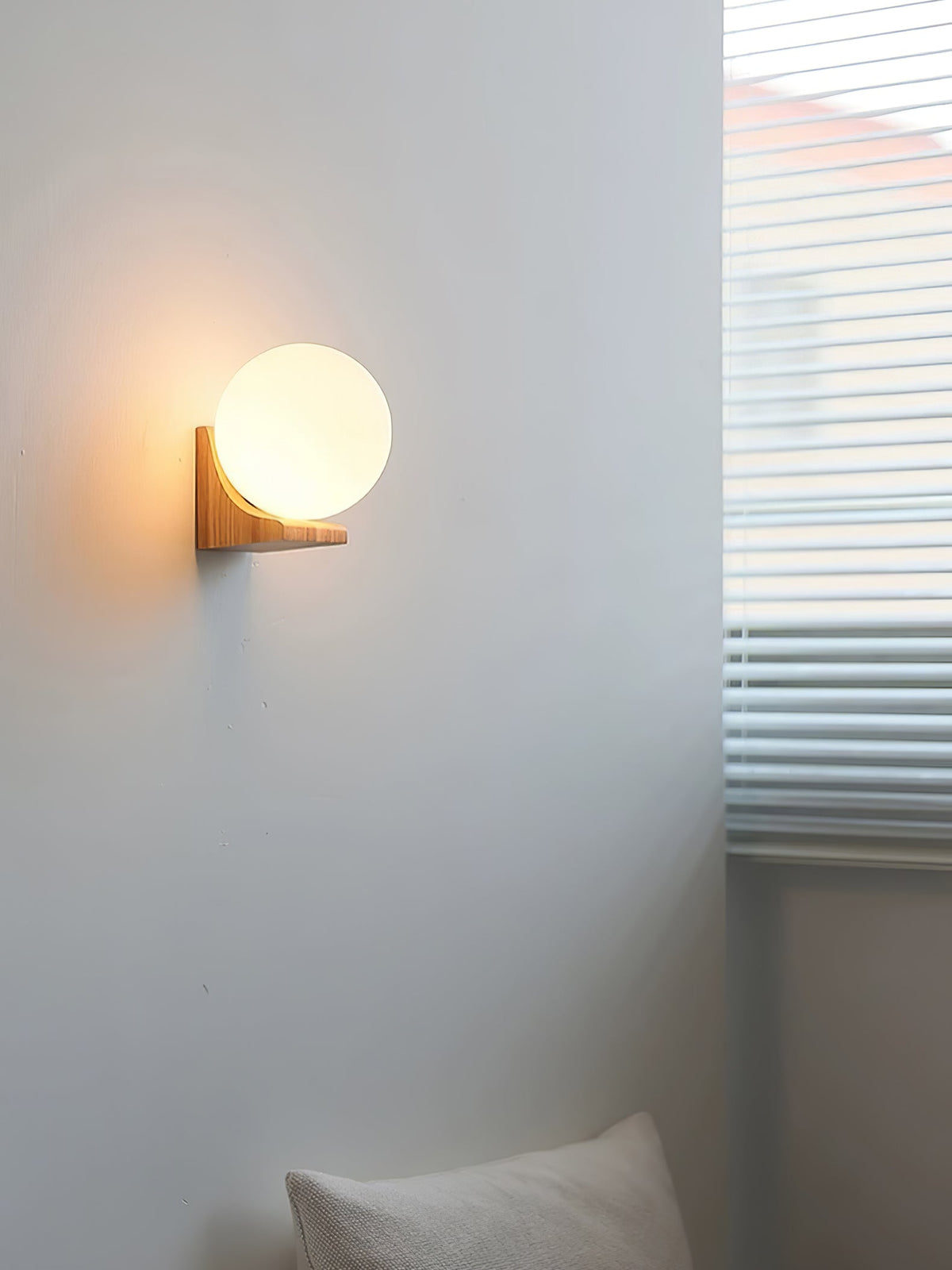 Tall room view of a Clérachic orb wall light glowing beside a window with blinds, with a cushion in the corner below.
