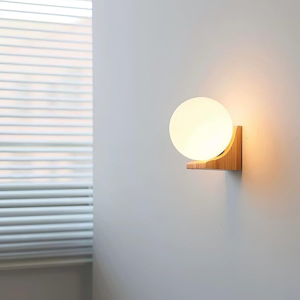 A Clérachic glass orb wall light glowing on a grey wall beside window blinds, its wooden bracket lit from the side.