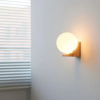 A Clérachic glass orb wall light glowing on a grey wall beside window blinds, its wooden bracket lit from the side.