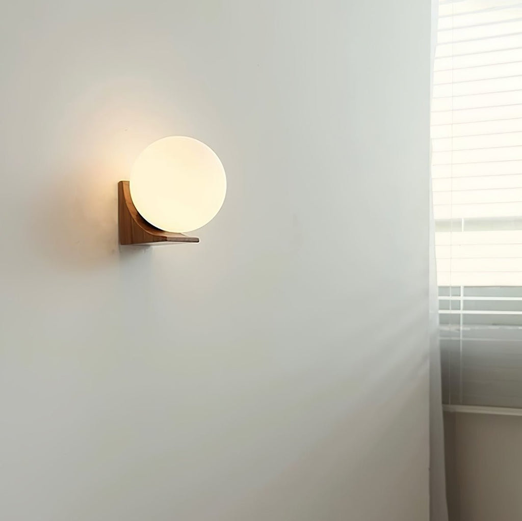 Lit Clérachic glass orb wall light in a light wood tone, mounted on a pale grey wall beside a curtain and blinds.