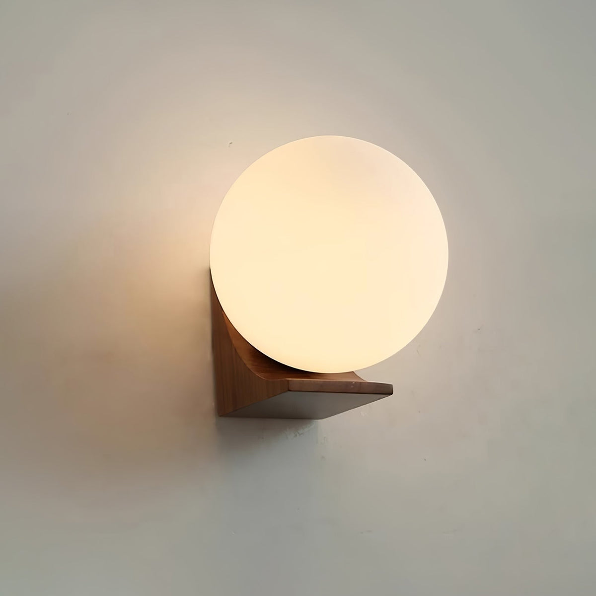 Lit Clérachic orb wall light in a walnut tone, its warm glow fading across a grey-green wall above the dark wood bracket.