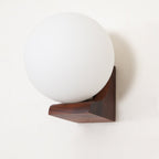 Close view of an unlit frosted glass orb on a dark walnut bracket, this Clérachic wall light against a cream wall.