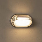 Clérachic white oval wall lamp with louvred slats over a frosted lens, lit warm on a rough plastered wall.