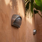 Clérachic close-up of a half-dome aluminium wall lamp with ribbed glass on a terracotta wall under palm leaves.