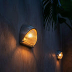 Clérachic half-dome aluminium wall lamp lit amber after dark on a courtyard wall, its ribbed glass glowing under a palm.