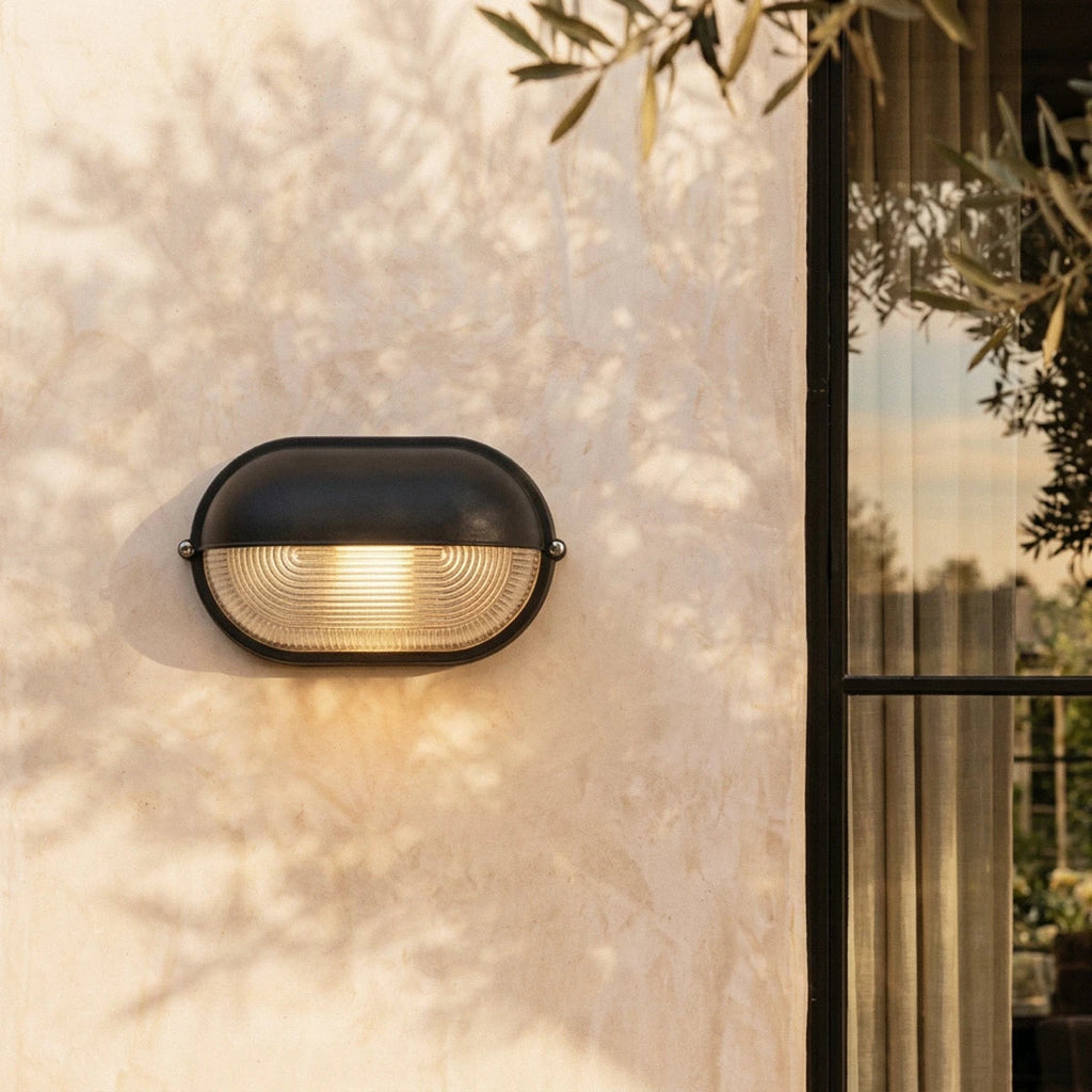 Clérachic black oval wall lamp with a hooded cap and ribbed glass, lit warm on a limewash wall with olive-leaf shadows.