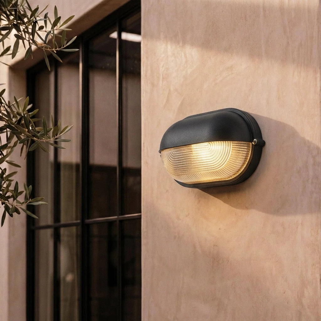 Clérachic black oval wall lamp lit beside a black-framed window on a blush plaster wall, with olive branches in view.