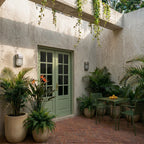 Clérachic upright galvanised wall lamps flanking a sage green French door in a plastered courtyard with palms and a bistro set.